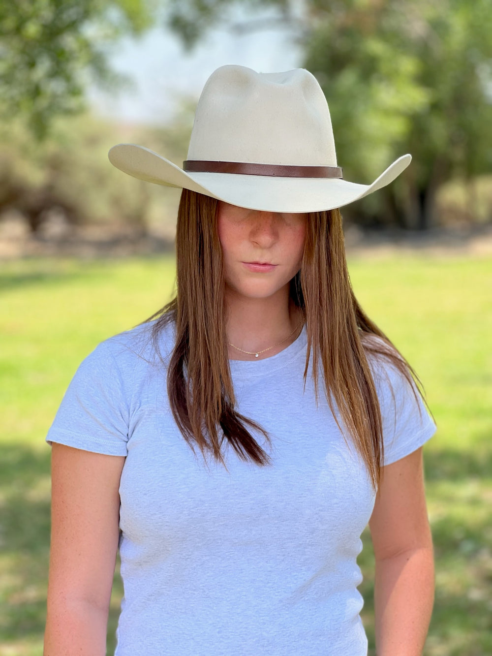 Person wearing a white cowboy hat and gray shirt outdoors with greenery in the background